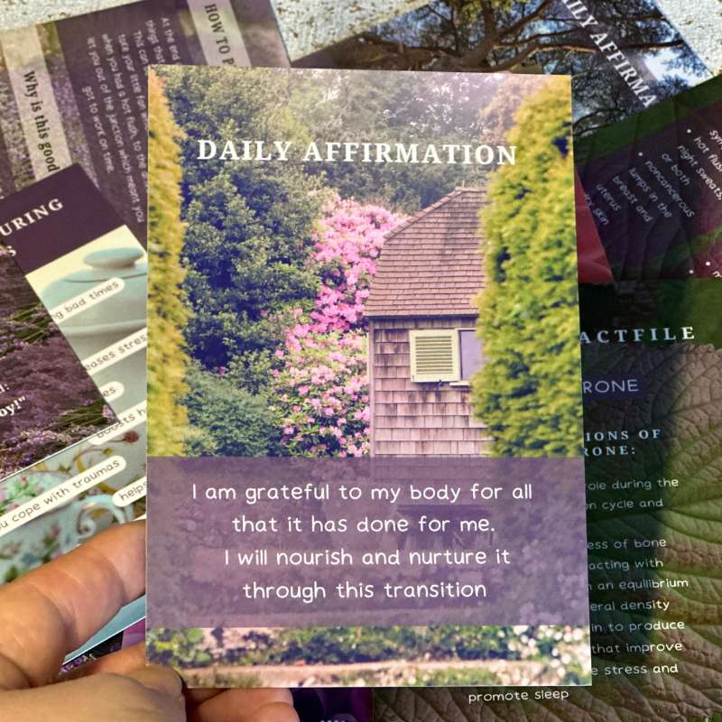 The deck of 50 better menopause cards lying next to the outer box, which is a brown Kraft box with a purple background sticker talking about the deck of cards. It is leaning against a vase.