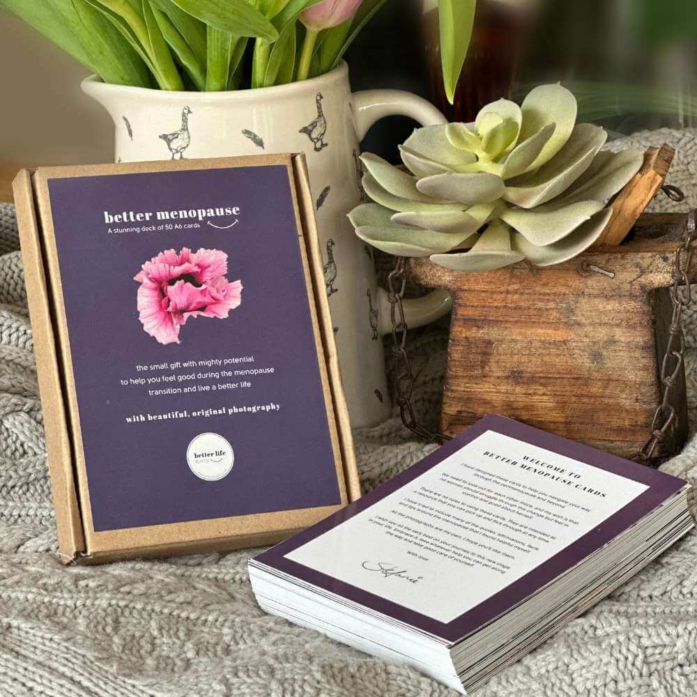 The deck of 50 better menopause cards lying next to the outer box, which is a brown Kraft box with a purple background sticker talking about the deck of cards. It is leaning against a vase.